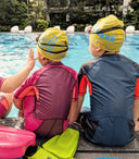 Two children in swim caps sitting by a pool with swim gear around them -Customised & Personalised Swimming Lessons for Toddlers in Singapore