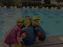 Three children wearing swim caps sitting by a pool listening to swim coach debrief - Learn to Swim Program in SIngapore for Young Children