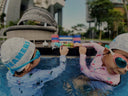 Two children in swim caps and goggles playing in a pool with a modern building in the background - Learn to Swim for Toddlers & Children in Singapore - Private Condo Swim Coaching