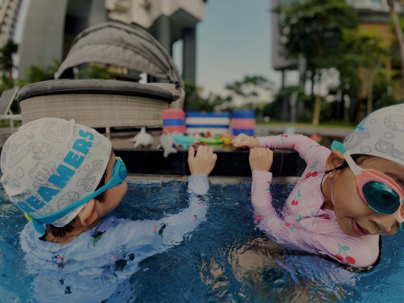 Two children in swim caps and goggles playing in a pool with a modern building in the background - Learn to Swim for Toddlers & Children in Singapore - Private Condo Swim Coaching